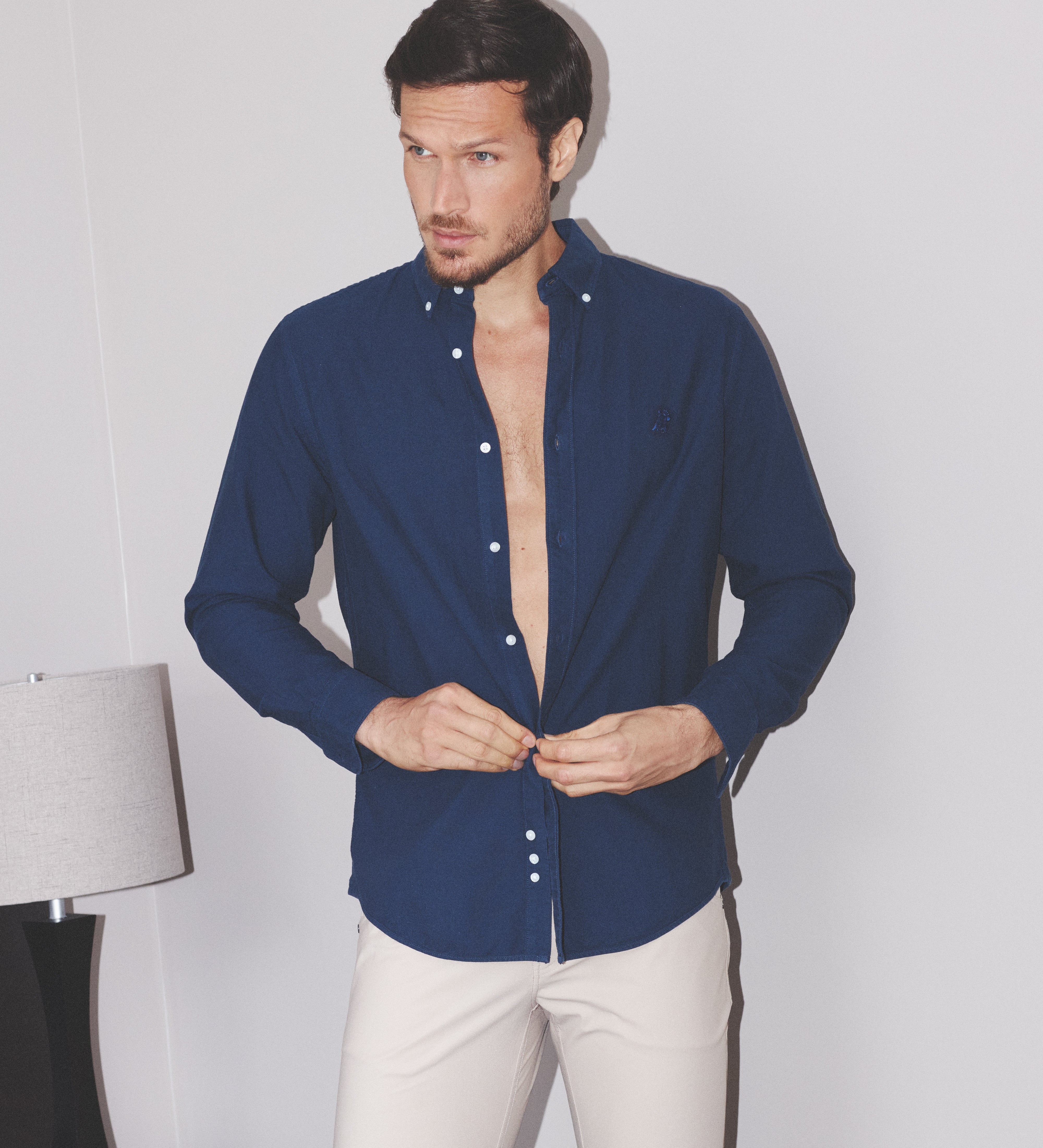 Man wearing a blue shirt with white buttons and beige pants against a plain background
