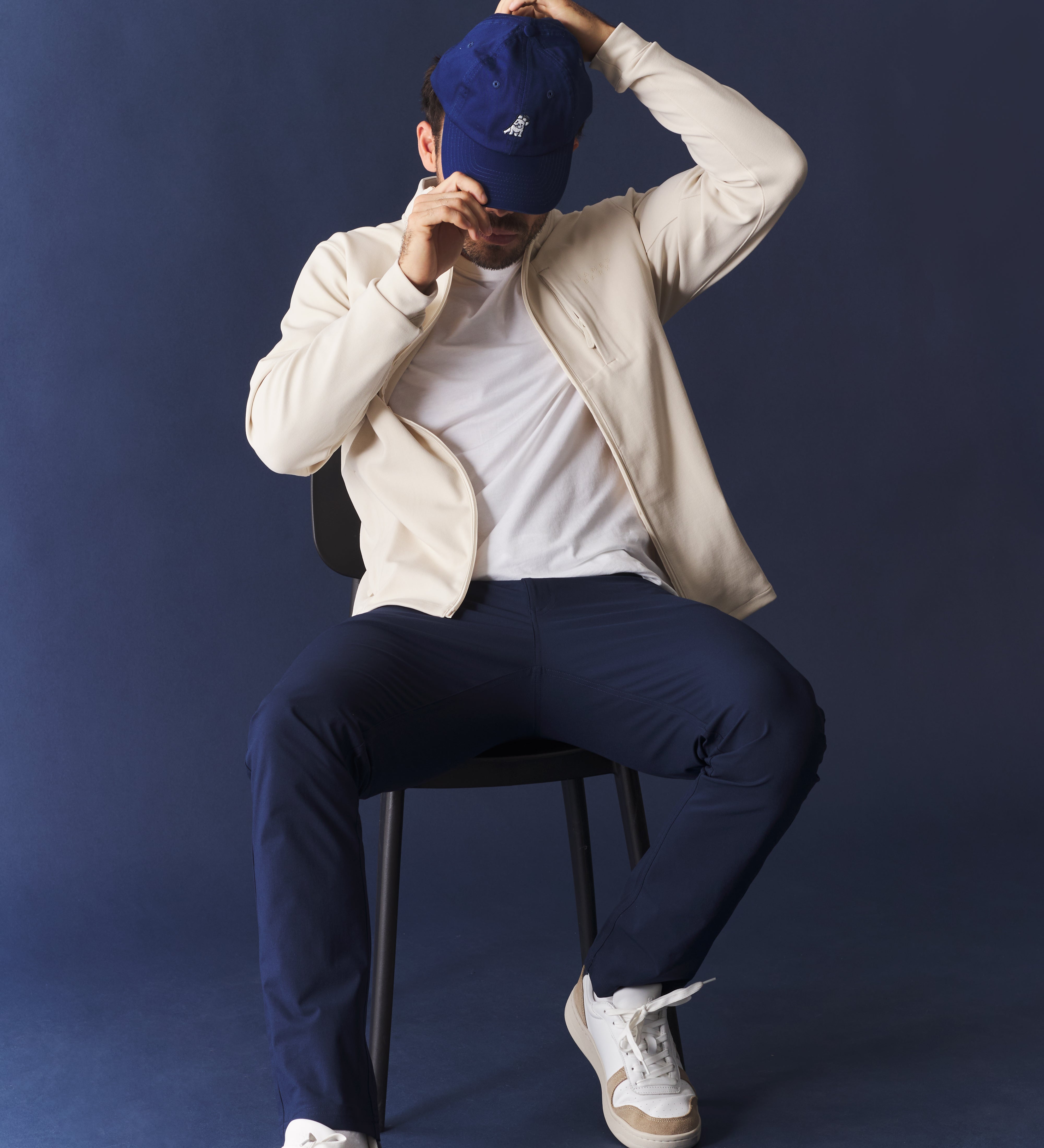 Person sitting on a chair wearing a white jacket, white shirt, navy pants, and white sneakers against a dark blue background.