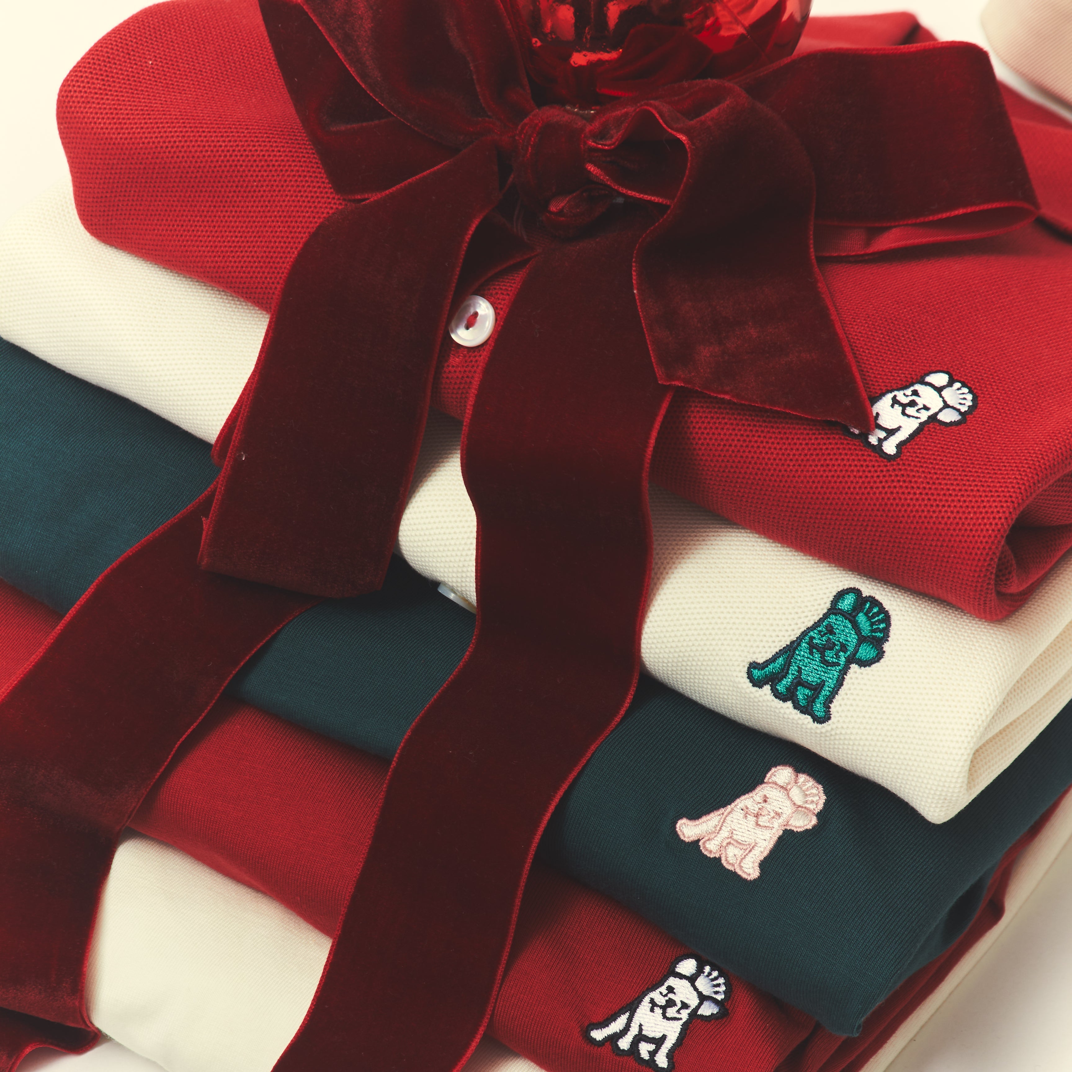 A stack of folded red and cream knit garments tied with a large dark red velvet ribbon, each with small embroidered dog motifs in different colors.