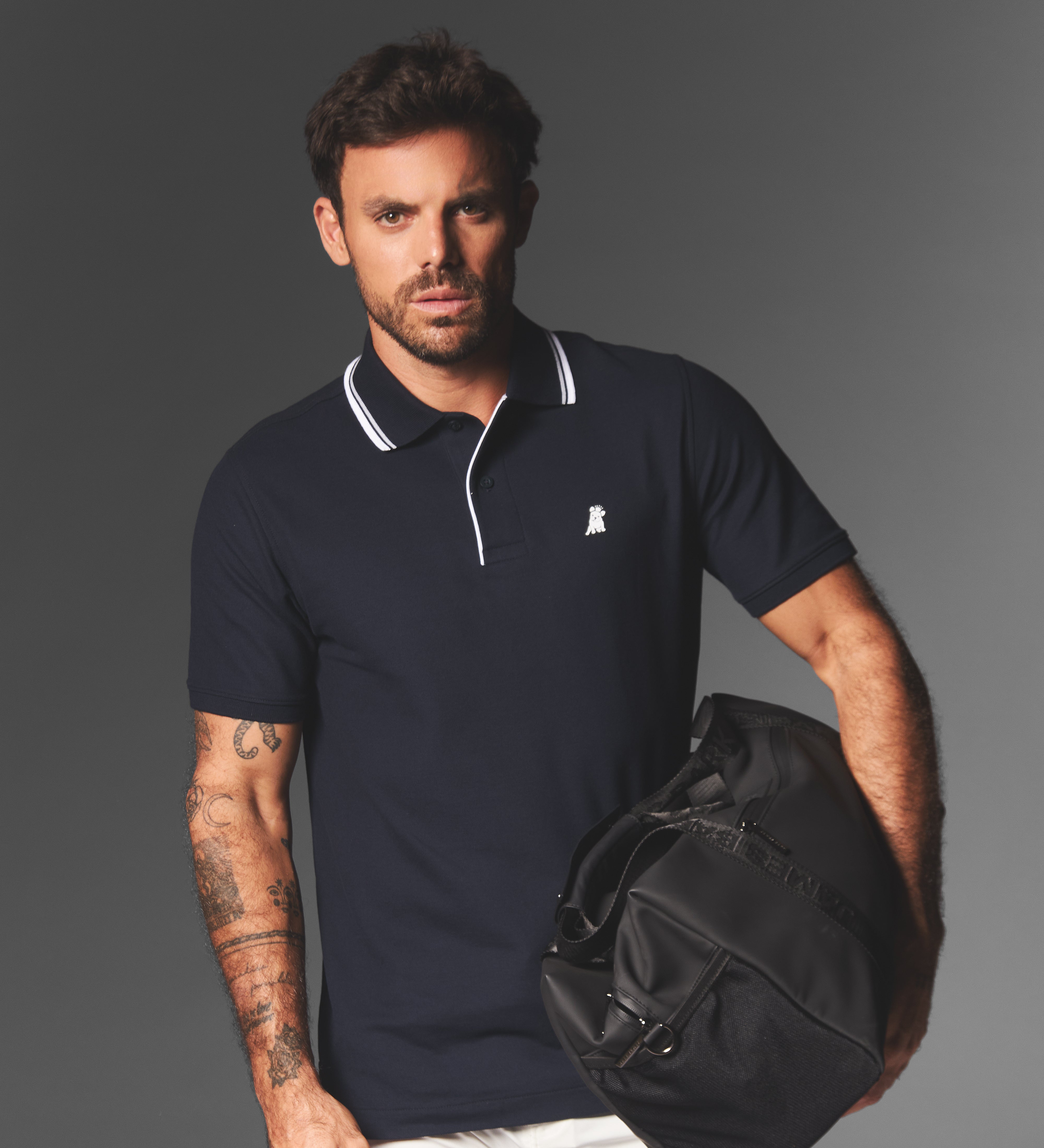 Man holding a black duffel bag wearing a navy polo shirt with a logo on a gray background