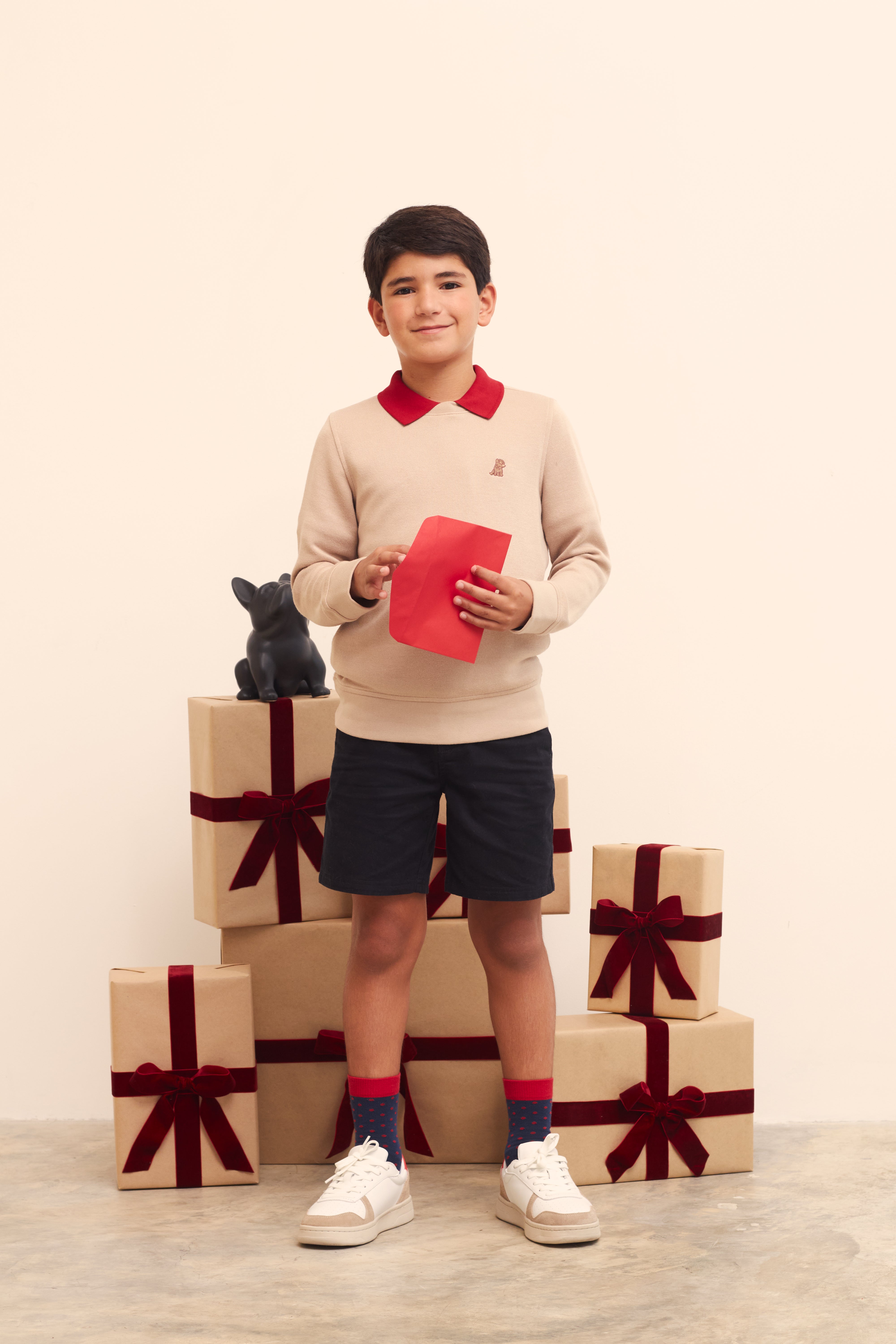 Young boy holding a red card with gift boxes and a small dog figurine on a beige background