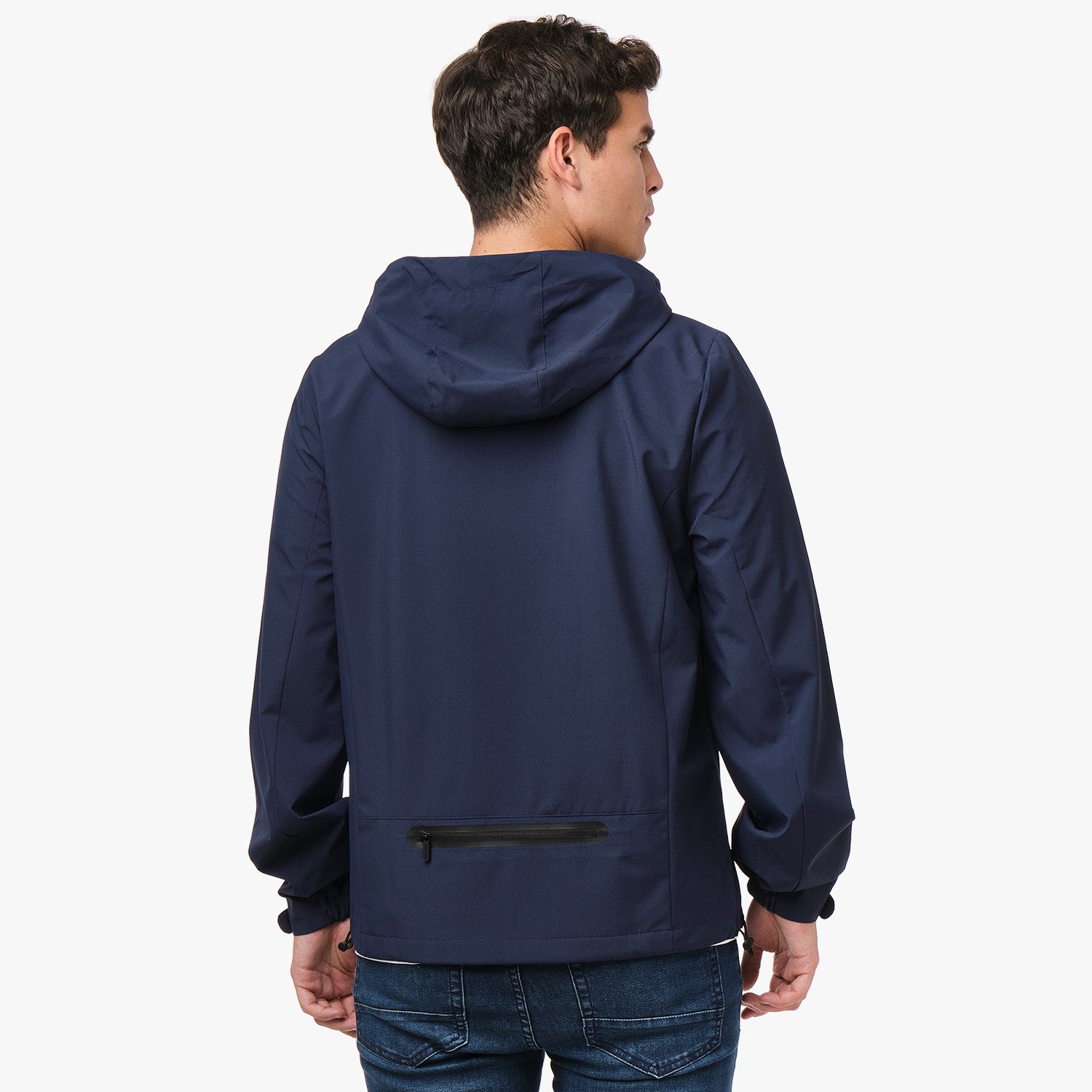 A man seen from the back wearing a navy blue hooded jacket with a small zippered pocket near the lower back. The jacket has long sleeves with elastic cuffs and is paired with blue jeans. The man is standing with his head turned slightly to the side, showing the profile of his face.