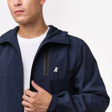 A man wearing a navy blue jacket with a hood, over a plain white t-shirt. The jacket features a black, waterproof zipper on the chest pocket and a small embroidered white dog with a crown on the left side of the chest. The man is slightly adjusting the front of the jacket with one hand, while his face is partially visible.