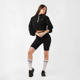 A model poses with her hands together, wearing a cropped jacket and sporty shorts, looking away from the camera against a neutral background.