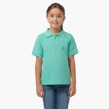 A young girl wearing a mint green polo shirt with a small embroidered logo on the chest, standing with her hands at her sides and smiling.