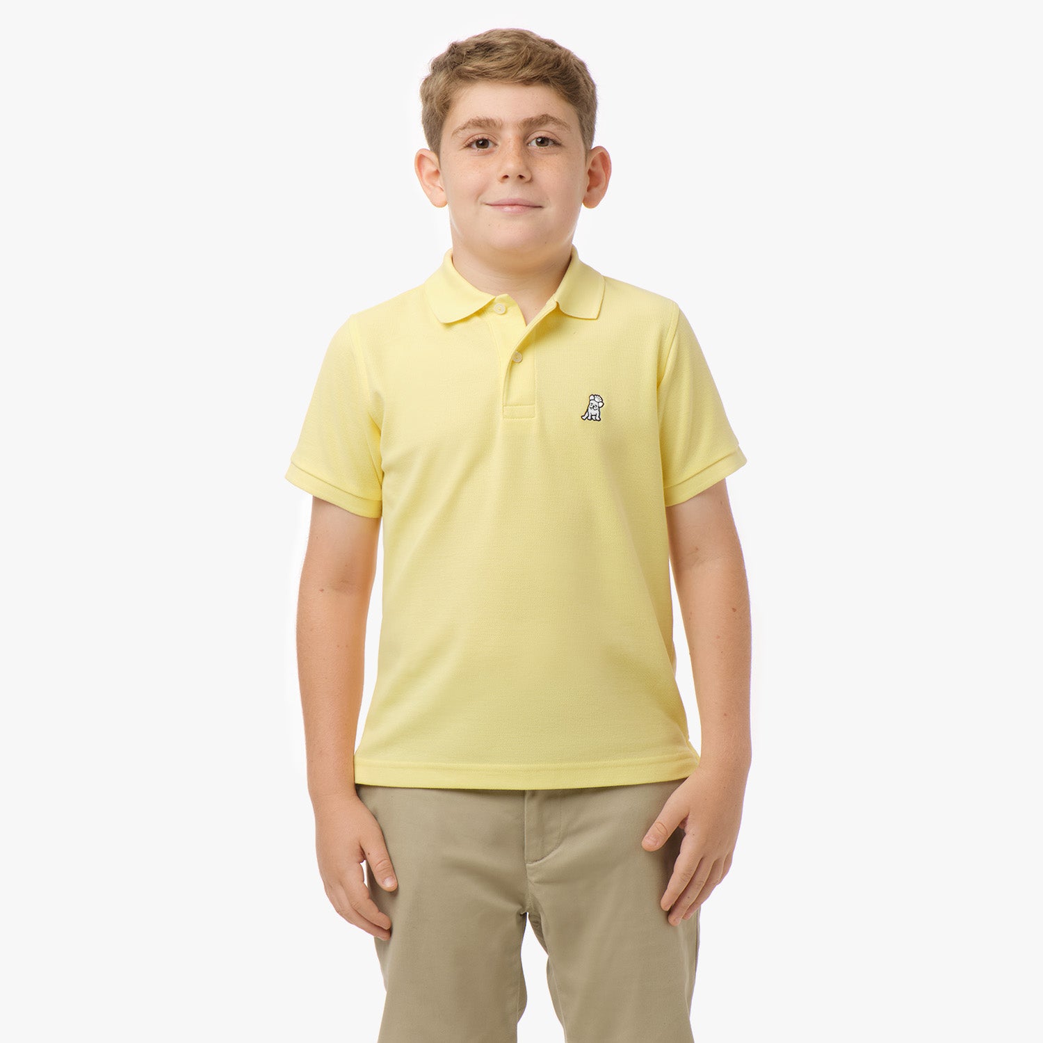 A young boy wearing a yellow polo shirt with a small embroidered logo on the chest, standing with his hands in his pockets and smiling, paired with beige pants.