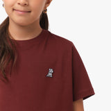 Close-up front view of a girl wearing the maroon t-shirt with a small white embroidered logo on the chest.