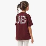 Rear view of a girl with braids wearing a maroon t-shirt with large "JB" lettering on the back, filled with a white pattern.