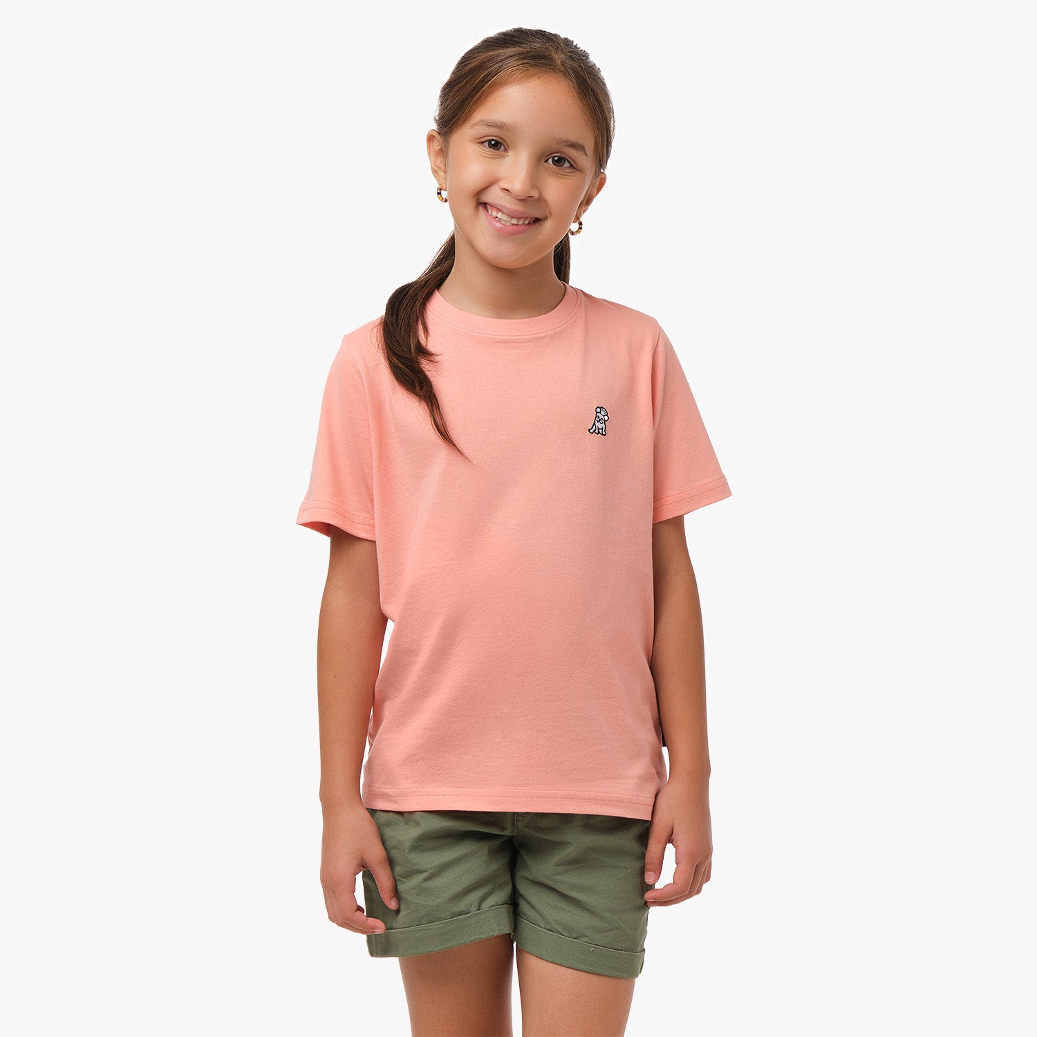 Front view of a girl wearing a light pink t-shirt with a small white embroidered logo on the chest, paired with olive green shorts, smiling with her hands by her sides.