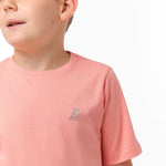 Close-up front view of a boy wearing the light pink t-shirt with a small white embroidered logo on the chest.