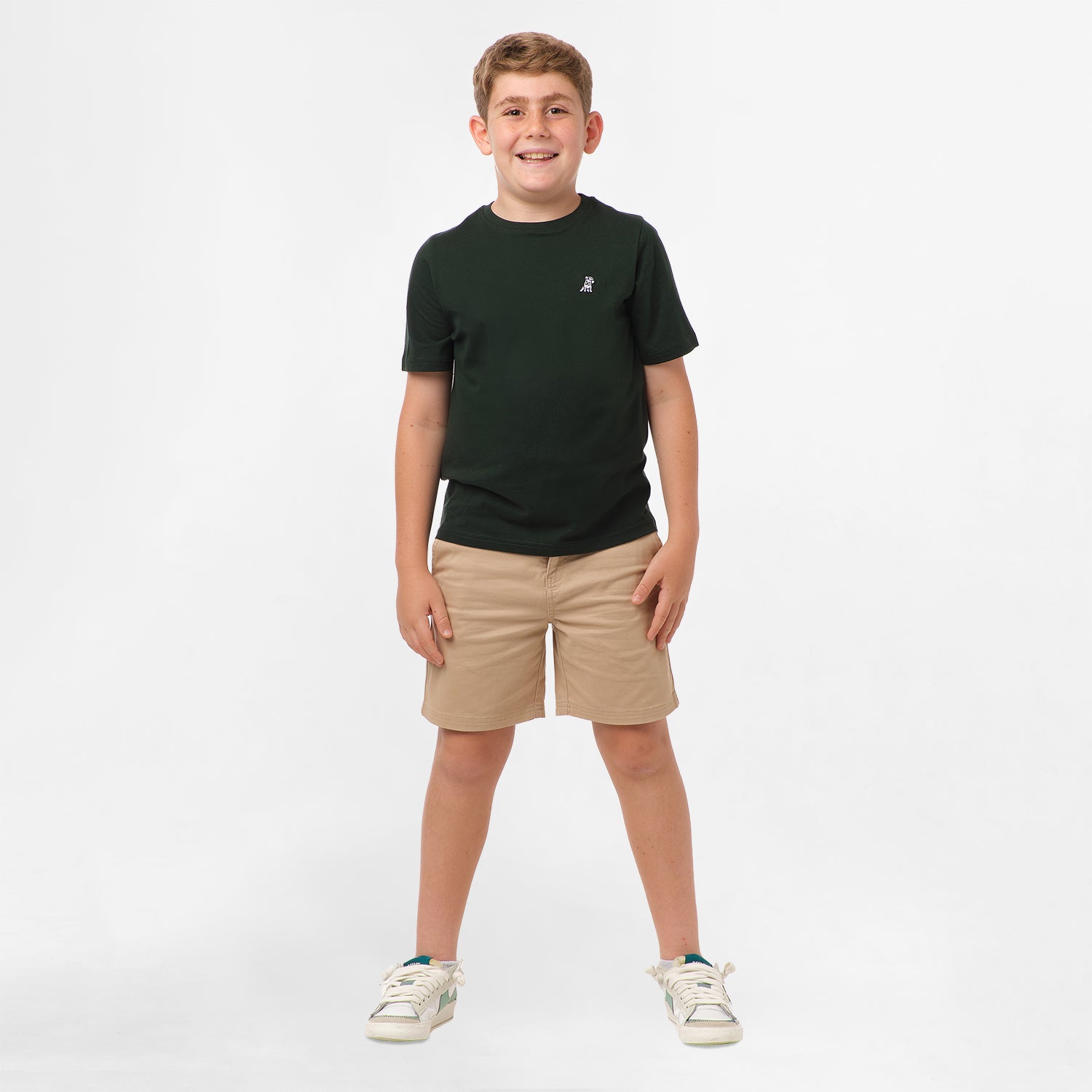 Full-body view of a boy wearing the black t-shirt and beige shorts, standing with his hands in his pockets and legs slightly apart.