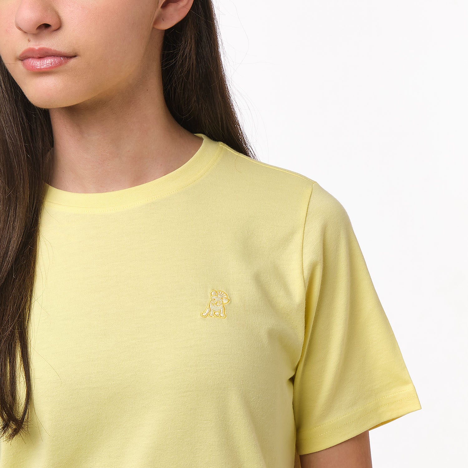 Close-up front view of a girl wearing the light yellow t-shirt with a small embroidered logo on the chest.