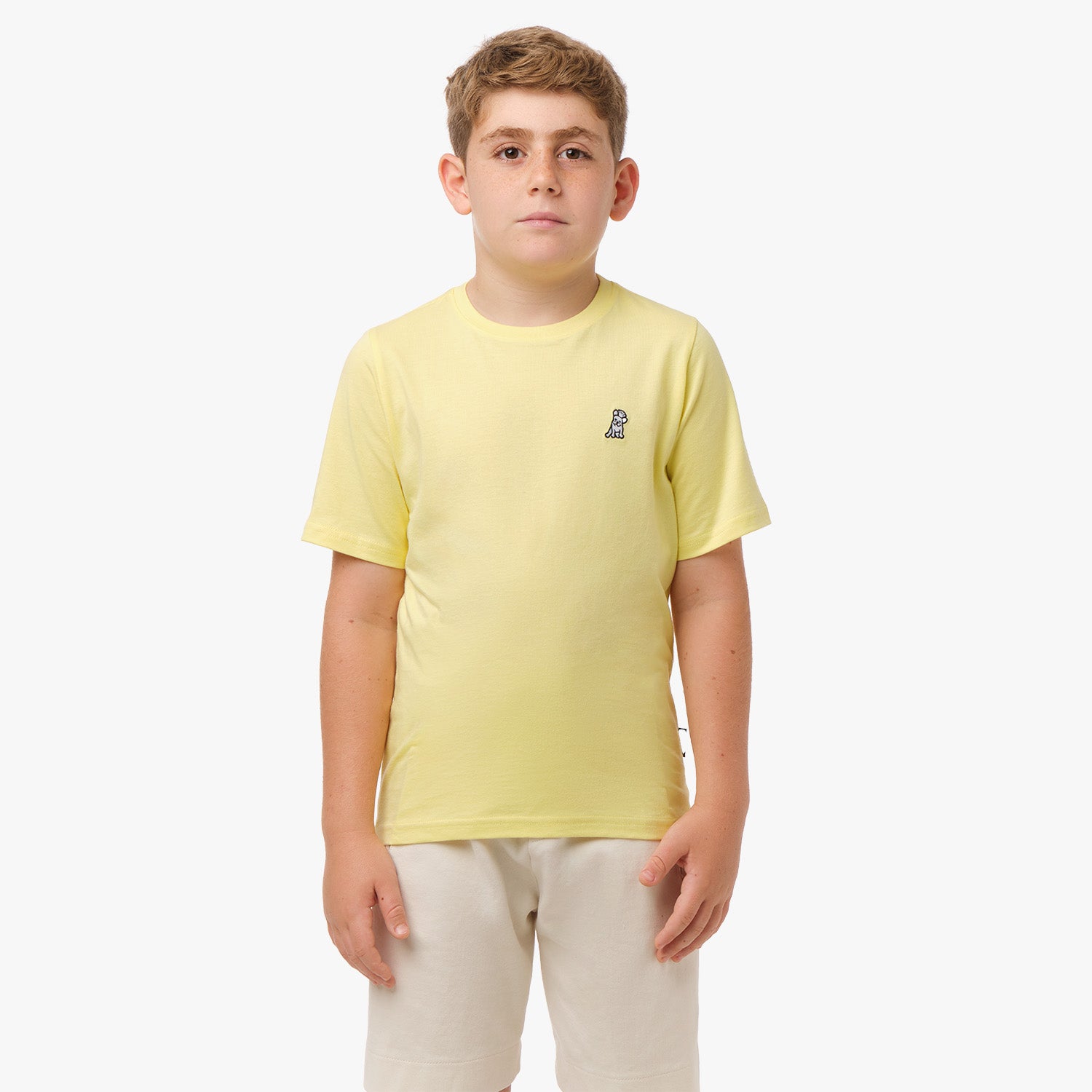 Front view of a boy wearing a light yellow t-shirt with a small white embroidered logo on the chest, paired with light beige shorts, standing with his hands in his pockets.