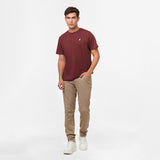 A full-body shot of the man in the maroon t-shirt and beige pants, standing casually with his hands in his pockets.