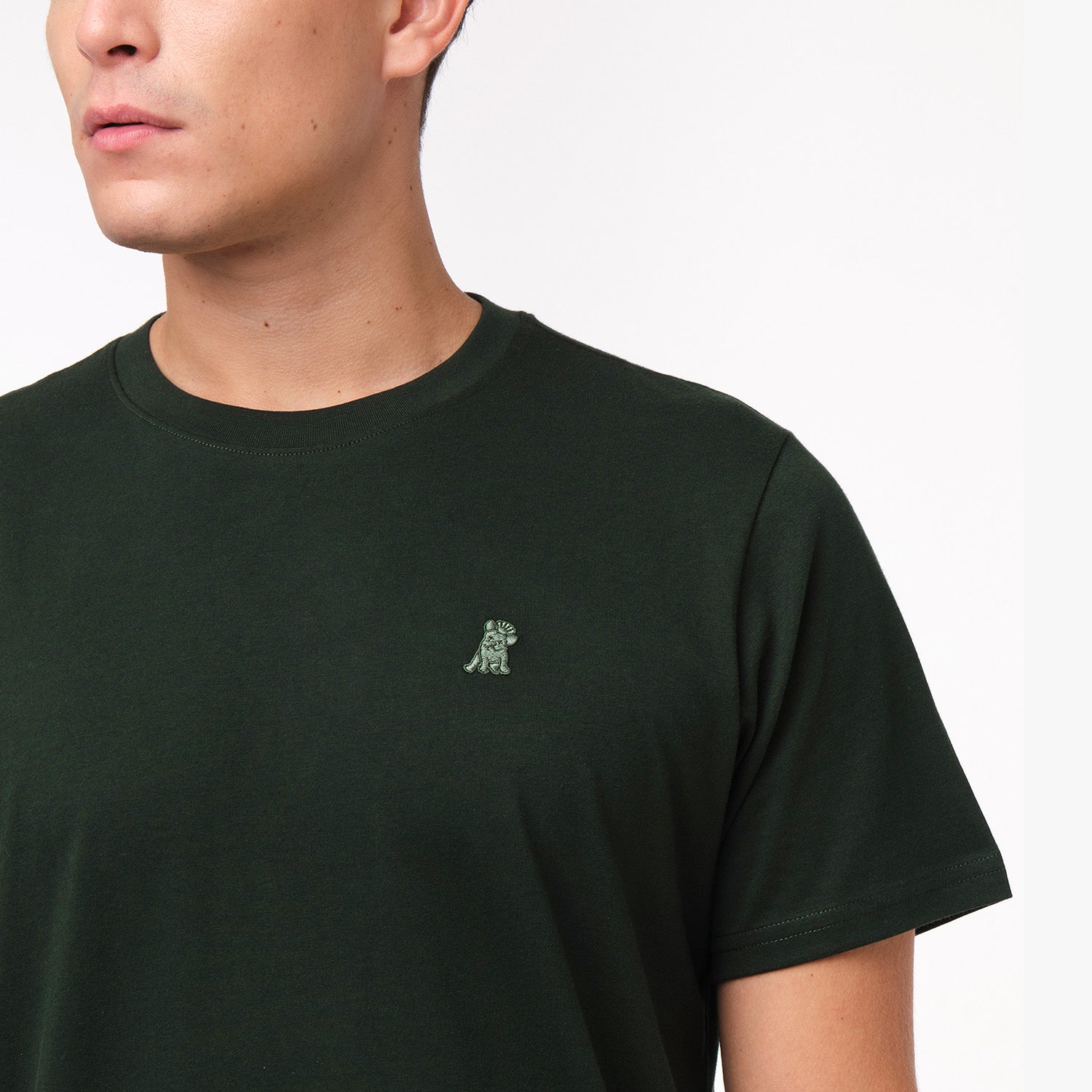 Close-up of the upper chest area of the dark green t-shirt, focusing on the small embroidered animal logo near the neckline.