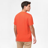 Back view of the man wearing the orange t-shirt, standing with his arms at his sides, paired with beige pants.