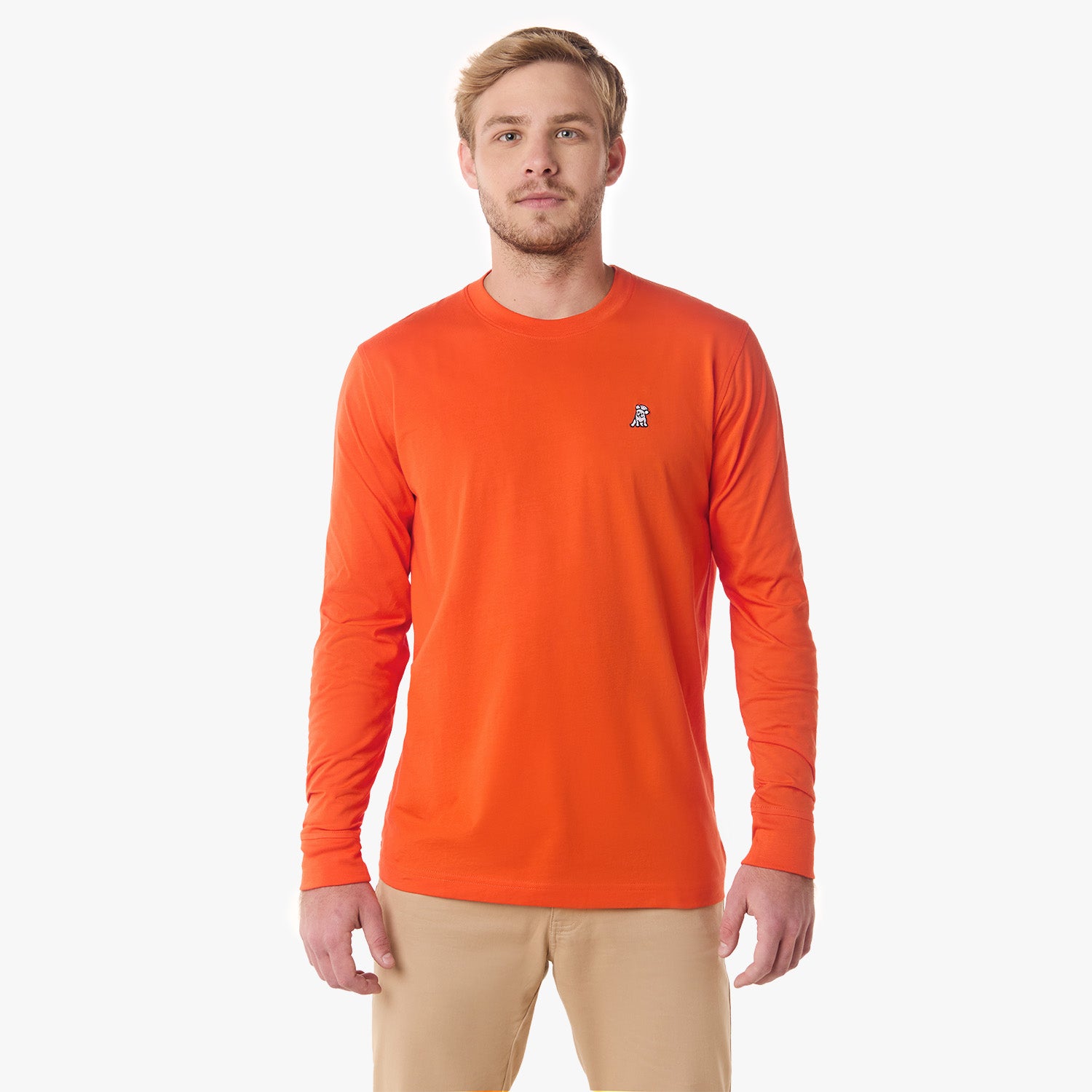 A man with a beard standing, wearing a long-sleeved bright orange sweater, looking forward, paired with beige pants.