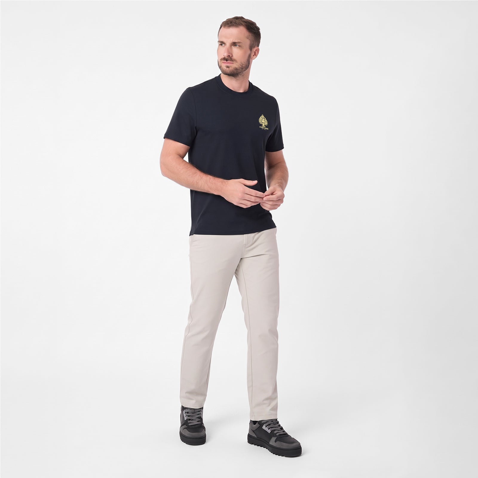 A man modeling a dark navy blue T-shirt with a small yellow logo on the left chest, paired with beige trousers and black sneakers