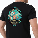 A man wearing a black T-shirt with a large, colorful vintage-style graphic on the chest. The graphic features a teal and orange shield design with sailboats