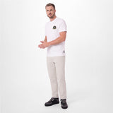 A man modeling a white T-shirt with a small circular graphic on the left chest, paired with beige trousers and black sneakers