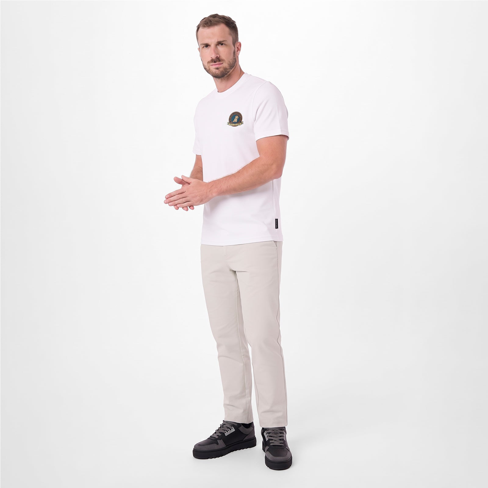 A man modeling a white T-shirt with a small circular graphic on the left chest, paired with beige trousers and black sneakers
