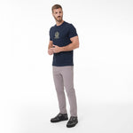 A man modeling a navy blue T-shirt with a gold crest logo on the left chest, paired with light gray trousers and black sneakers
