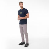 A man modeling a navy blue T-shirt with a gold crest logo on the left chest, paired with light gray trousers and black sneakers