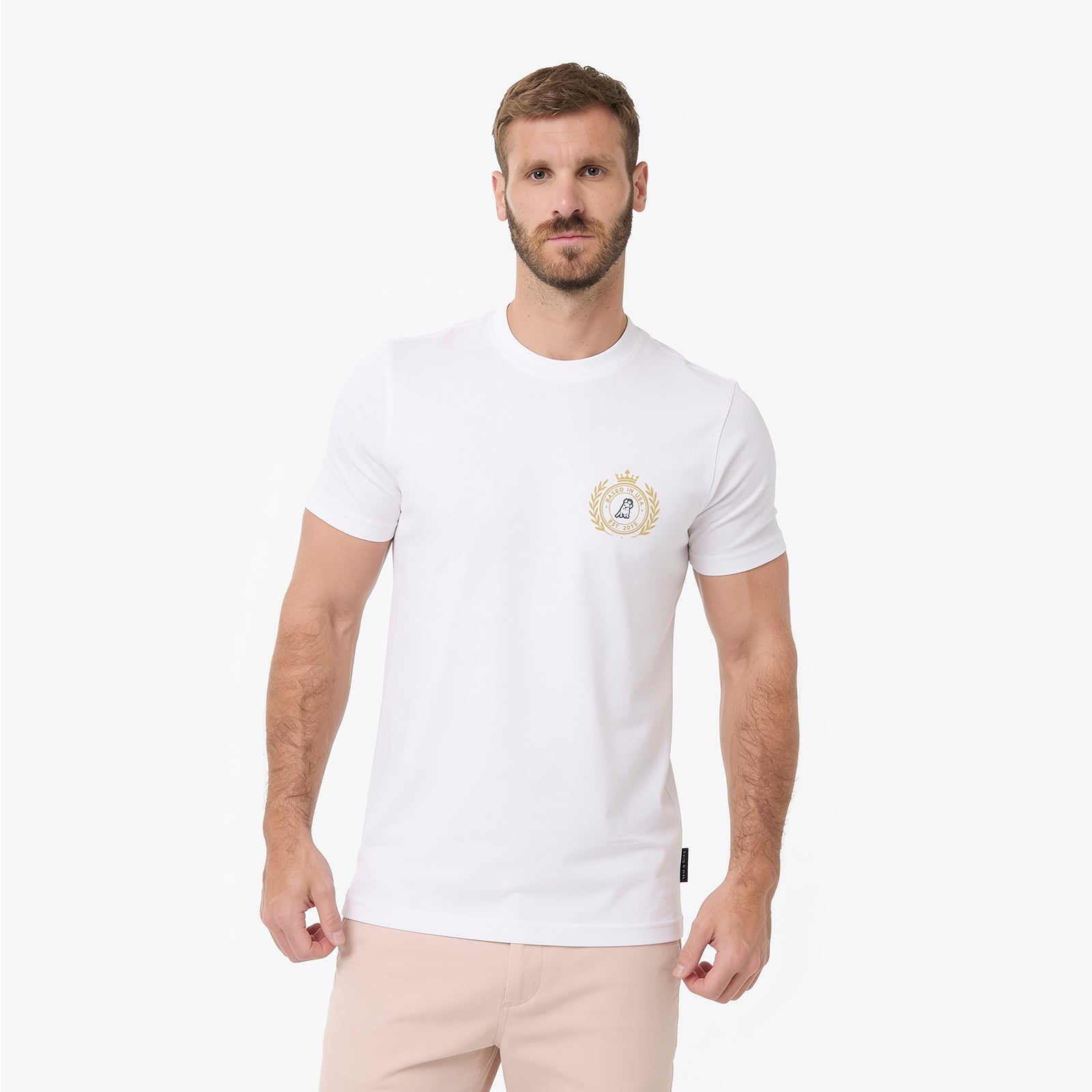 A man wearing a white T-shirt with a small gold and blue crest logo on the left chest