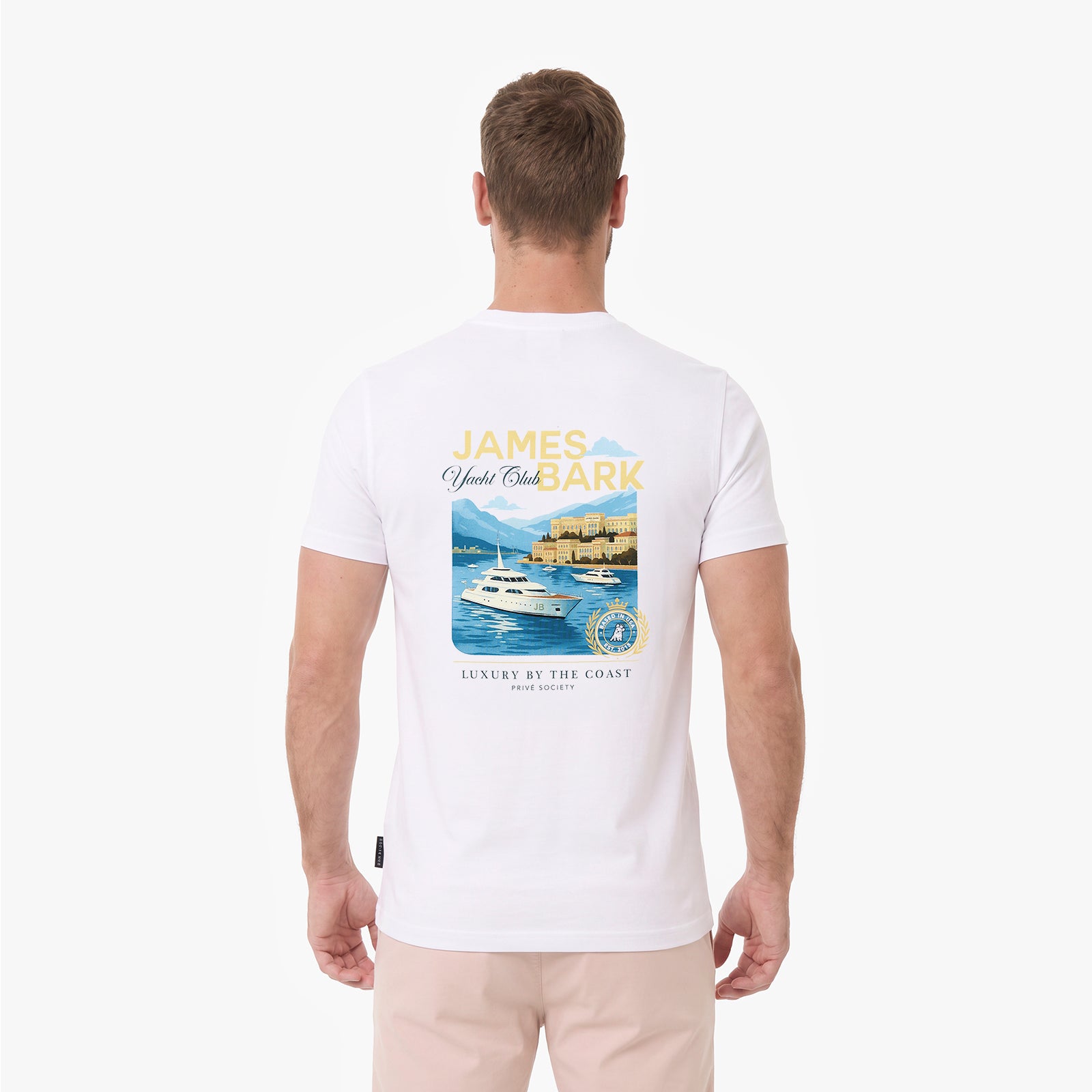 A man viewed from behind wearing a white T-shirt with a large graphic print on the back. The graphic features the text 'JAMES BARK' in yellow and blue, above an illustration of a yacht on blue water with mountains and a lighthouse.