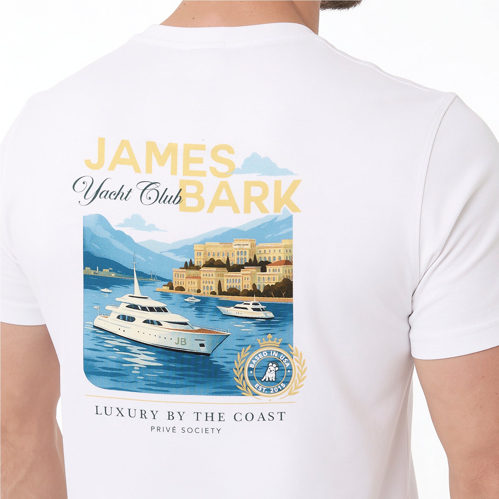 A man wearing a white T-shirt, showing the back of the shirt with a large graphic print. The graphic features the text 'JAMES Yacht Club BARK' in yellow and black, an illustration of yachts on blue water with mountains and a villa in the background, and a gold crest seal.