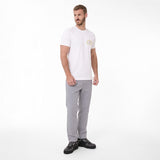 A man modeling a white T-shirt with a small circular gold and blue logo on the left chest, paired with light gray trousers and black sneakers
