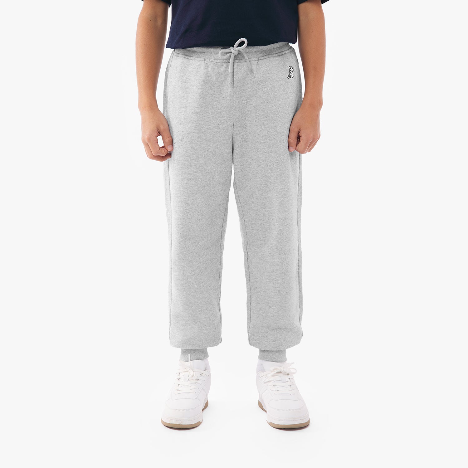 A person wearing light gray sweatpants with a drawstring waistband, a small embroidered logo near the pocket, and white socks. They are paired with green and white sneakers. The person is standing upright with arms relaxed by their sides, wearing a black top, and the background is plain white.