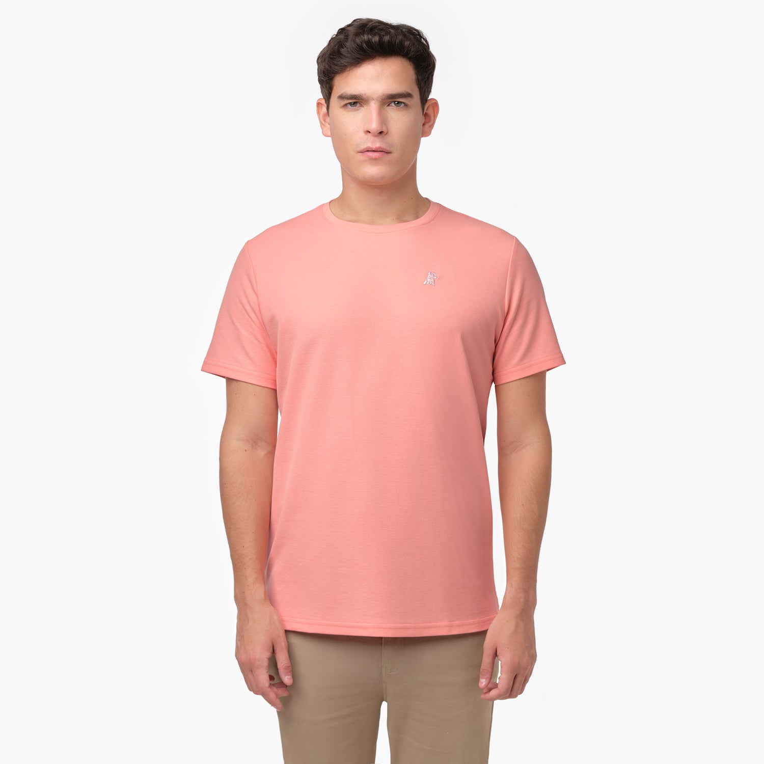 Male model showcasing the front of a peach/coral men's crew neck t-shirt with a logo, paired with tan chino pants, shot on white.