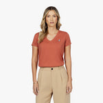 Front waist-up view of a female model wearing a rust-colored v-neck t-shirt with a small white embroidered logo on the left chest, paired with beige trousers
