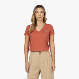 Front waist-up view of a female model wearing a rust-colored v-neck t-shirt with a small white embroidered logo on the left chest, paired with beige trousers