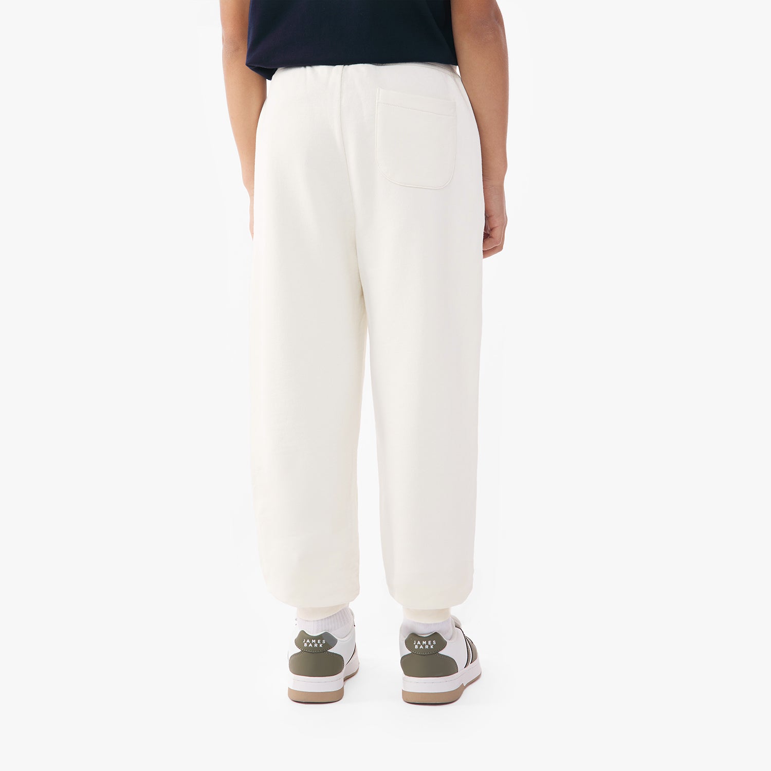 Back view of a child wearing off-white jogger pants with a single back pocket, paired with green and white sneakers and a green top.