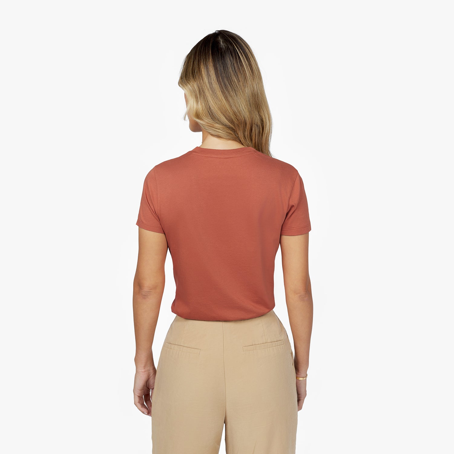 Back waist-up view of a female model wearing a plain rust-colored short-sleeve t-shirt tucked into beige trousers