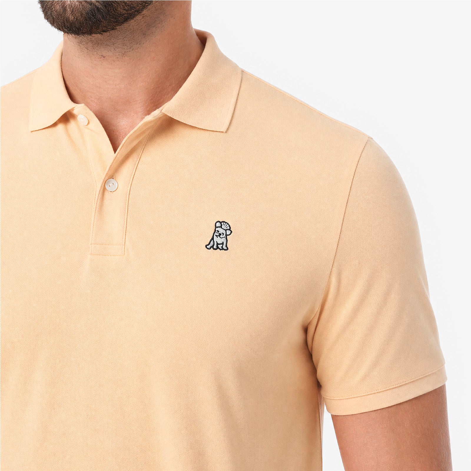 A close-up of a man wearing a light beige polo shirt with a small embroidered dog logo on the chest