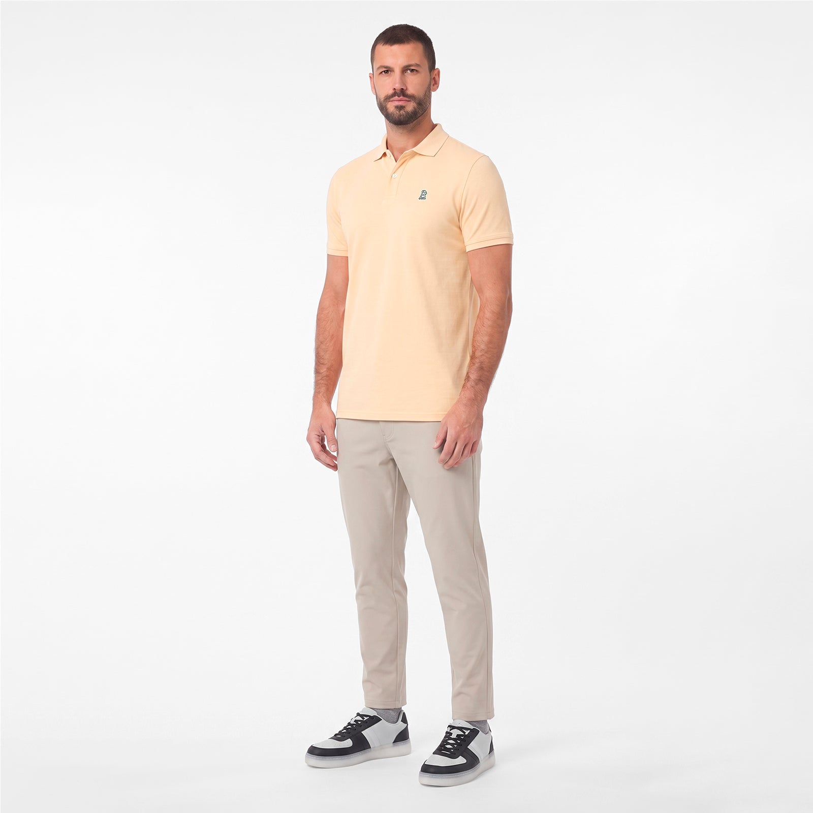 A man modeling a pale yellow polo shirt with beige trousers and black-and-white sneakers, standing against a plain white studio background.