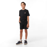 Full body shot of a boy smiling and standing, wearing black chino shorts, a black t-shirt with a logo, white socks, and navy sneakers. He has one hand in his shorts pocket