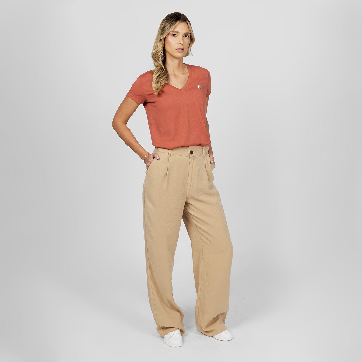 Full-length front view of a female model wearing a rust-colored v-neck t-shirt with a white logo, wide-leg beige trousers, and white sneakers, standing against a light grey background