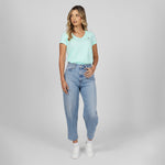 Full-length angled view of a female model wearing a mint green v-neck t-shirt with a white logo, light wash jeans, and white sneakers, looking slightly to the side against a light grey background