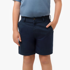 Close-up front view, waist-down, of a boy wearing navy blue chino shorts with a dusty blue t-shirt. One hand is in the front side pocket. The shorts feature a button closure and belt loops