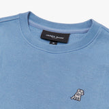 A close-up of a light blue crewneck sweatshirt, highlighting the ribbed collar and fabric texture. The black neck label displays the brand name "JAMES BARK" with "Est. 2015" beneath it, and a size "M" tag hanging below. On the chest, there is a small embroidered logo of a smiling dog waving. The fabric appears soft and finely knit.