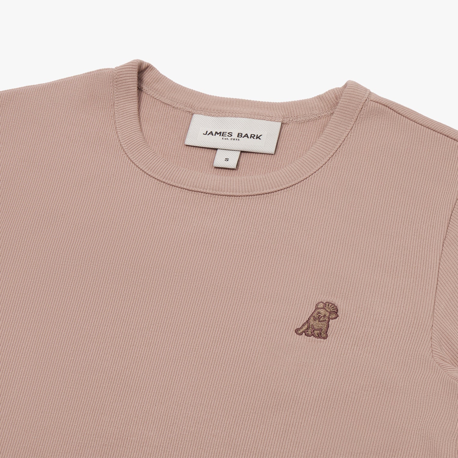 A close-up of a dusty rose-colored short-sleeve T-shirt, showing the ribbed crewneck collar and finely knit fabric texture. The white neck label displays the brand name "JAMES BARK" with "Est. 2015" beneath it, along with the size "S." A small embroidered logo of a dog in a darker brown thread is positioned on the left chest area. The material appears soft and high-quality.