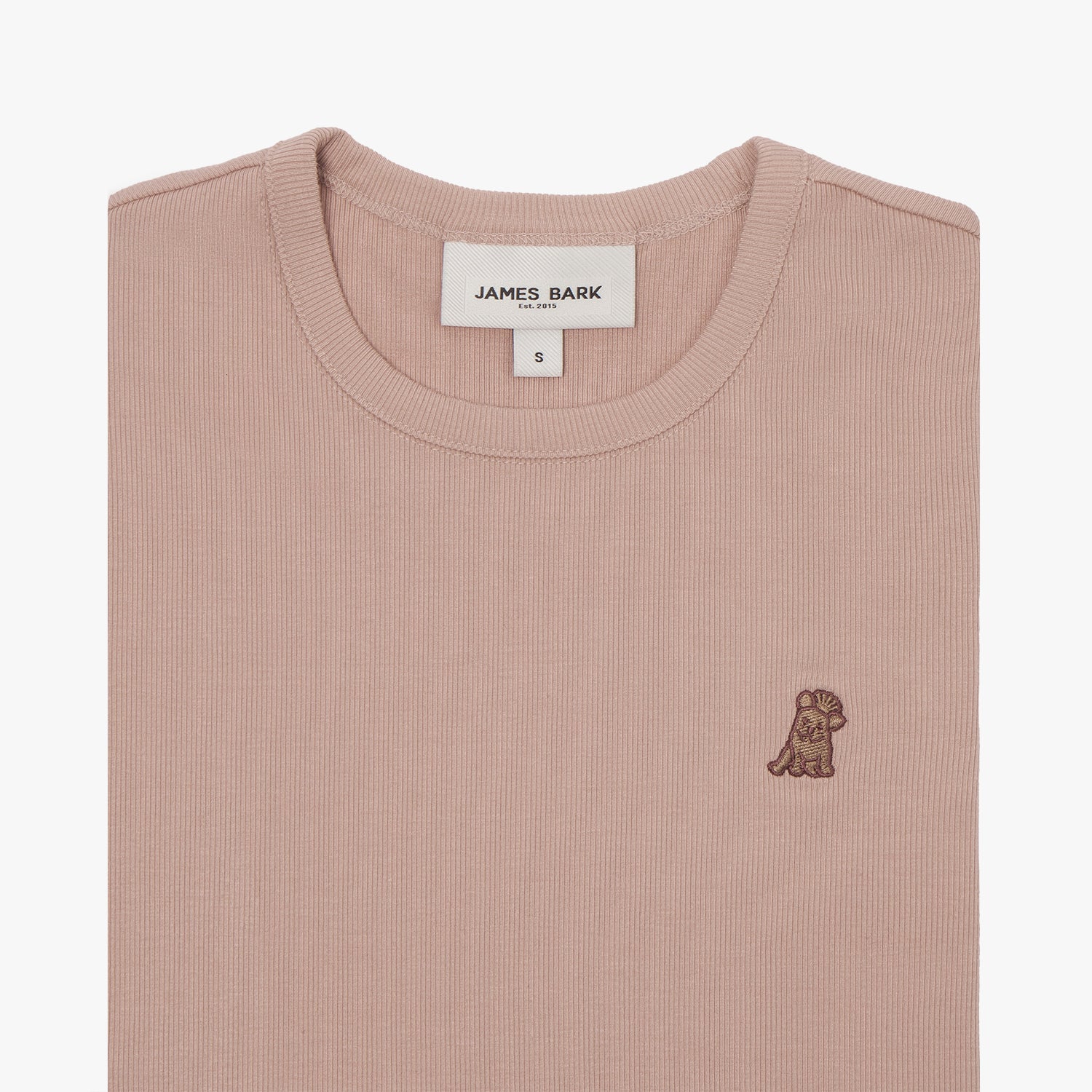 A close-up of a dusty rose-colored short-sleeve T-shirt, neatly folded to showcase the ribbed crewneck collar and finely knit fabric texture. The white neck label displays the brand name "JAMES BARK" with "Est. 2015" beneath it, along with the size "S." A small embroidered logo of a dog in a darker brown thread is positioned on the left chest area. The material appears soft and high-quality.