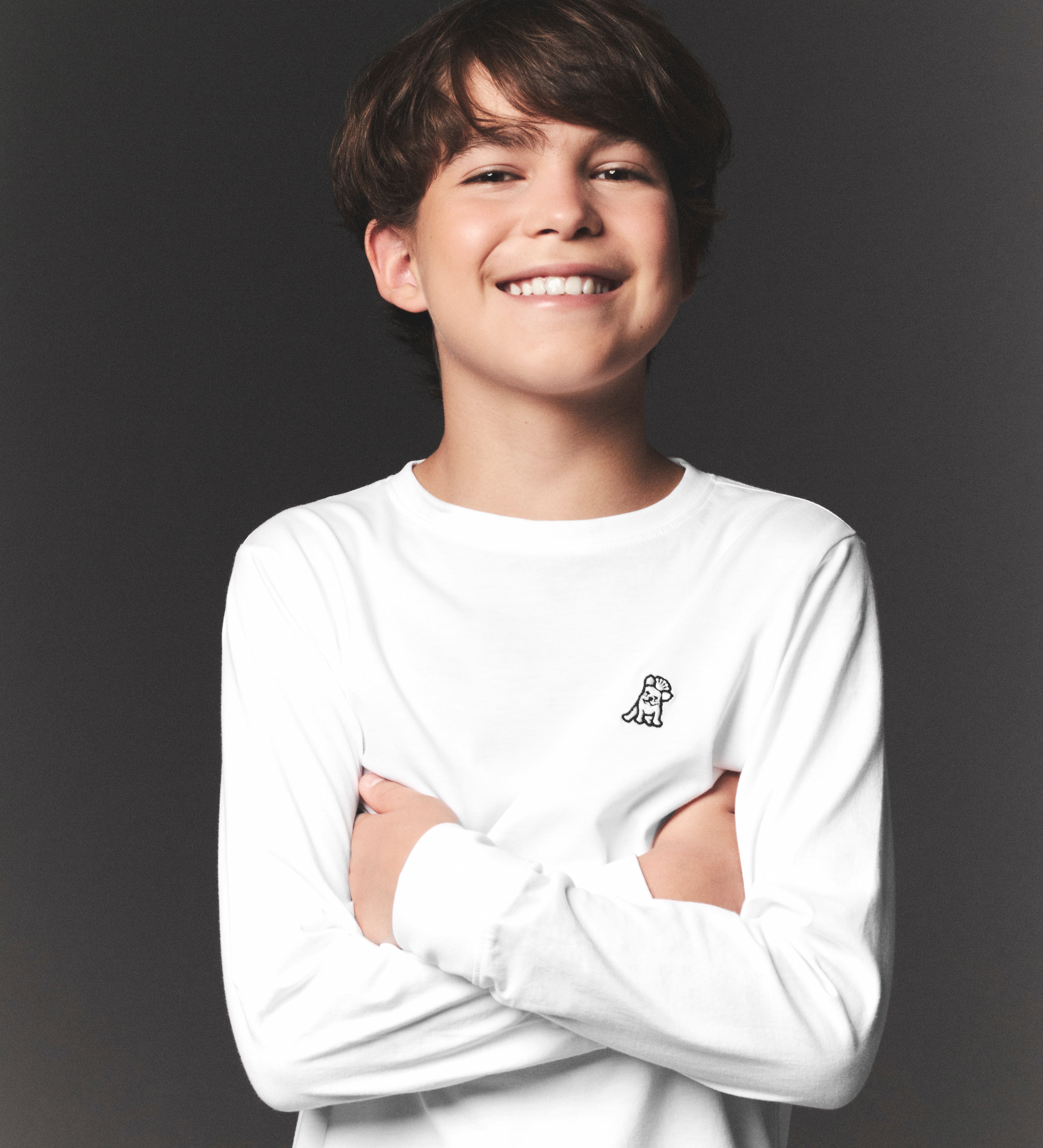 Young boy wearing a white long-sleeve shirt with a logo on a dark background