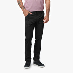 A close-up of a man wearing black pants, paired with a light pink shirt. The image focuses on the front of the pants, showing a slim, tailored fit and hands in pockets.