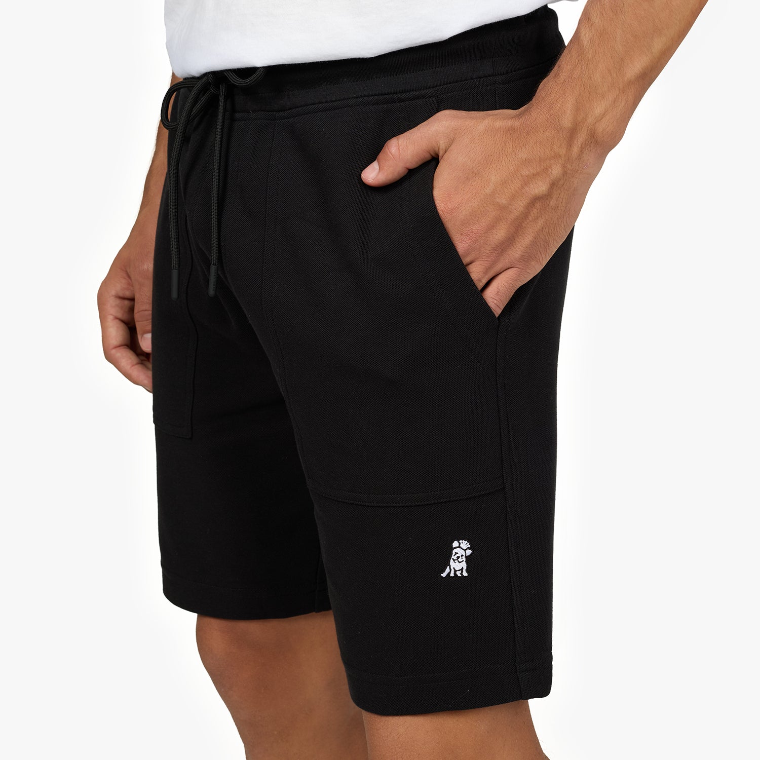 A close-up image of a man wearing black shorts with a drawstring waist and side pockets. The shorts have a small embroidered white dog logo on the lower left leg. The man's right hand is casually placed in his pocket, and he is wearing a white t-shirt. The background is plain white.