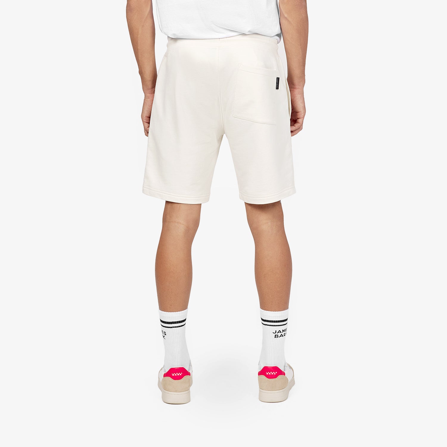 This image showcases the back view of the off-white casual shorts. The model wears a simple white t-shirt with the shorts, which have a patch pocket on the back with a small tag. The look is completed with white socks featuring black stripes and sneakers with pink branding, adding a pop of color to the outfit